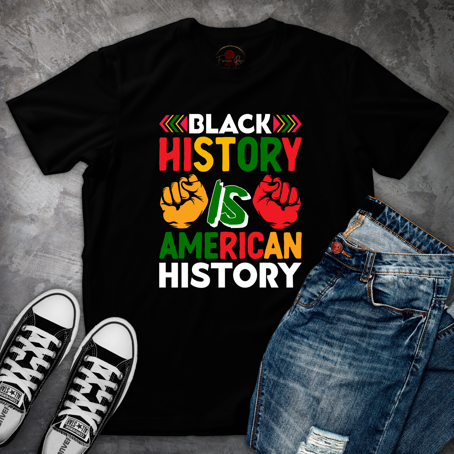 A black t-shirt with a graphic design that says "BLACK HISTORY IS AMERICAN HISTORY" in bold, colorful letters, along with two fist-pumping graphic elements. The t-shirt is placed on a gray surface, and there are a pair of jeans and black and white sneakers visible in the image.