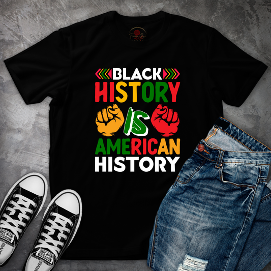 A black t-shirt with a graphic design that says "BLACK HISTORY IS AMERICAN HISTORY" in bold, colorful letters, along with two fist-pumping graphic elements. The t-shirt is placed on a gray surface, and there are a pair of jeans and black and white sneakers visible in the image.